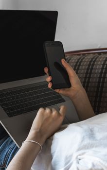 Woman with a laptop is laying on a sofa and using a smartphone. Study and work online, freelance. Self employed girl working with her notebook laying on a couch with a phone.