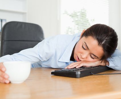 Frontal view of a beautiful woman sleeping on a keyboard while holding a cup of coffee