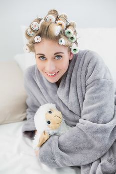 Happy relaxed blonde woman in hair curlers cuddling a plush sheep