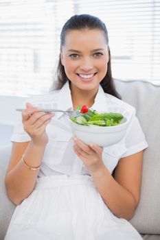 Smiling pretty woman eating healthy salad sitting on sofa