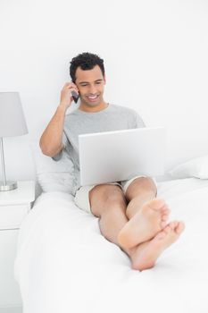 Casual smiling man using cellphone and laptop in bed