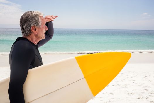 Senior man with surfboard shielding eyes at beach