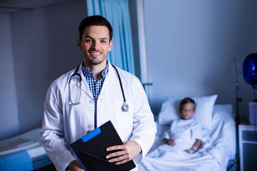 Portrait of male doctor standing with clipboard in ward at hospital