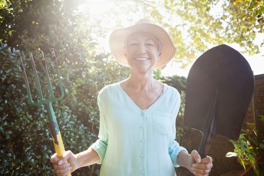 Back lit portrait of smiling senior woman holding garden fork and shovel