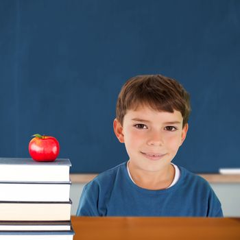Cute boy smiling against red apple on pile of books in classroom