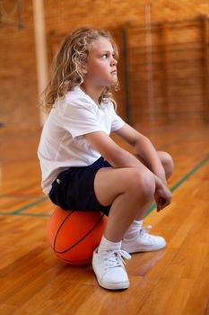Schoolgirl sitting on a basketball in basketball court