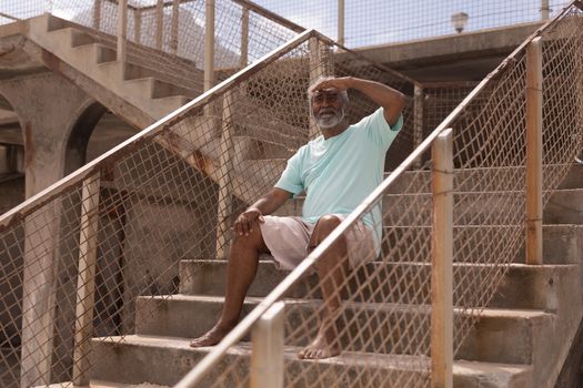 Senior man shielding eyes and sitting on promenade stairs at beach