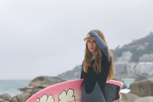 Woman standing with surfboard shielding eyes at beach
