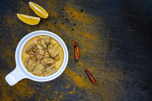 Orange spicy chicken curry in a simple white plate on a dark background. Around hot pepper, lemon, curry powder. Indian traditional cuisine. Flat lay. Top view, copy space.