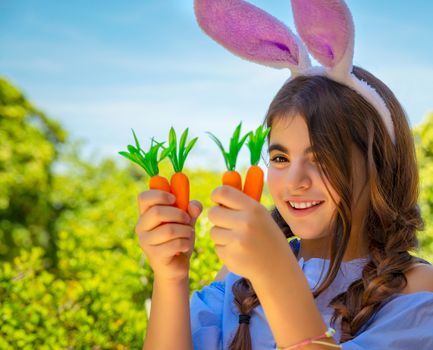 Portrait of a cute little bunny girl with carrots in hands enjoying Easter, having fun gardening in the yard on a spring sunny day, traditional outfit for Easter celebration, happy holiday
