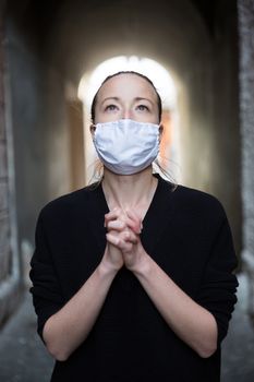 Coronavirus outbreak. Young caucasian woman wearing medical protection face mask praying over coronavirus global pandemic, for salvation of humanity, health, anxiety and depression reduction