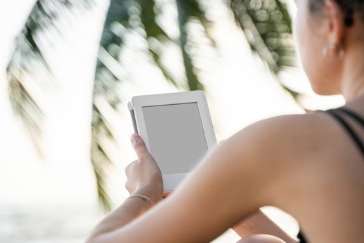 Woman holding e-reader outdoors by the sea with a palm trees on background. Relaxing and enjoying with a reading a favorite books outdoor in a travel