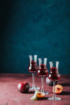Plums strong alcoholic drink in grappas wineglass with dew. Hard liquor, slivovica, plum brandy or plum vodka with ripe plums on dark blue and claret bordeaux concrete surface