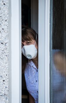 woman in a protective medical mask looking out from behind a partially open door. Coronavirus epidemic UK