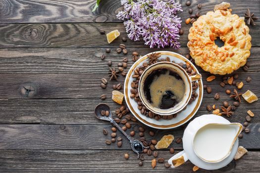 Rustic wooden background with cup of coffee, milk, peanut tart, sugar ginger and lilac flowers. White vintage dinnerware and spoon. Breakfast at summer morning. Top view, place for text.