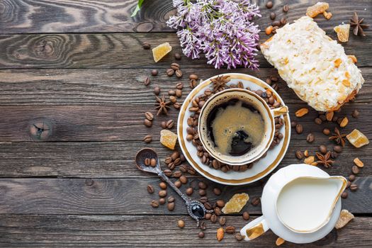 Rustic wooden background with cup of coffee, milk, bun with marzipan and lilac flowers. White vintage dinnerware and spoon. Breakfast at summer morning. Top view, place for text.