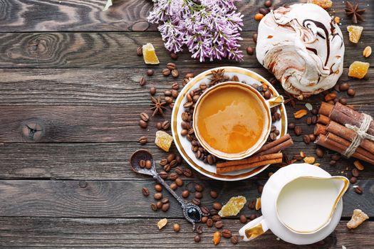 Rustic wooden background with cup of coffee, milk, meringue, cinnamon and lilac flowers. White vintage dinnerware and spoon. Breakfast at summer morning. Top view, place for text.