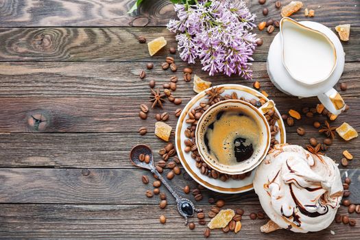 Rustic wooden background with cup of coffee, milk, meringue, cinnamon and lilac flowers. White vintage dinnerware and spoon. Breakfast at summer morning. Top view, place for text.