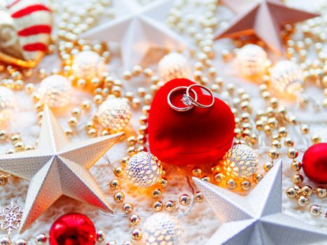 Christmas and New Year star decorations on white knitted background. Red heart gift box with wedding diamond rings, metal light bulbs with delicate pattern, golden beads.