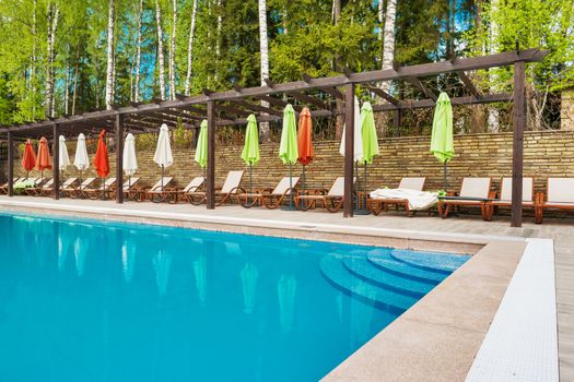 Sun loungers and parasols around the pool with clear blue water. Birch tree forest around pool.