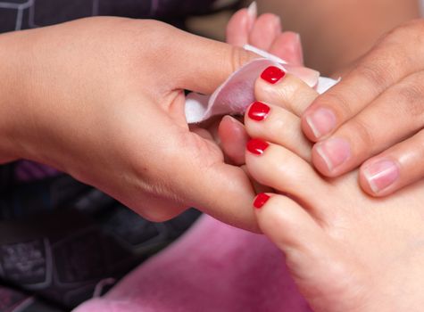 Woman receiving toenail pedicure service by pedicurist at nail salon. Beautician coating red gel color toenail to customer at nail and spa salon. Foot care and toenail treatment at nail salon. 