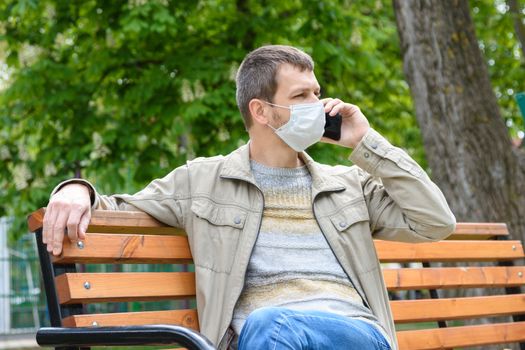 A man in a protective mask walks through the park and crouches on a bench talking on the phone