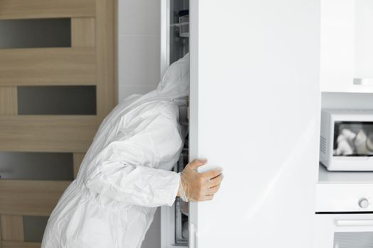 Man in white protective suit and in face medical mask stares in a refrigerator in the white kitchen at home during coronavirus. Stay at home. Enjoy cooking at home. COVID-19 global pandemic.