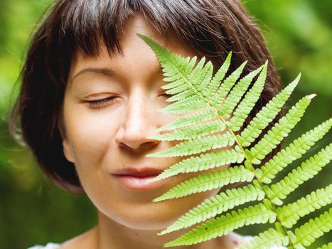 Young woman is hiding her eye with fern leaf. Symbol of life, tranquility and unity with nature. Summer in forest.