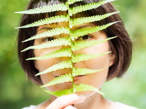 Young woman is hiding her eyes with fern leaf. Symbol of life, tranquility and unity with nature. Summer in forest.