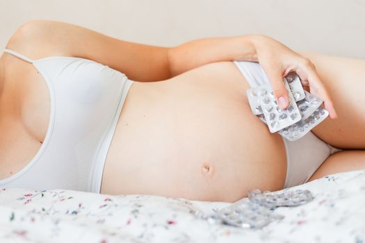 Pregnant woman in white underwear lying in bed with many pills. 