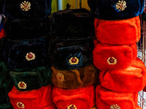 Colorful Russian winter hats with earflaps. Metal pin with red USSR star and Russian double-headed eagle symbols. Souvenir shop in Moscow, Russia.
