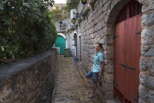 The young man stands near the wooden gate in the narrow street of the old town of Budva, Montenegro.