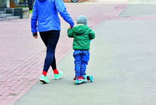 The kid in blue jeans and a green jacket on a children's scooter walks with his mother on the paved sidewalk.