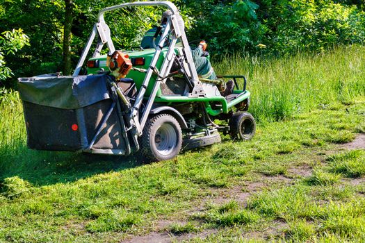 A professional tractor mower driven by a public utility worker climbs up the slope and mows tall grass.