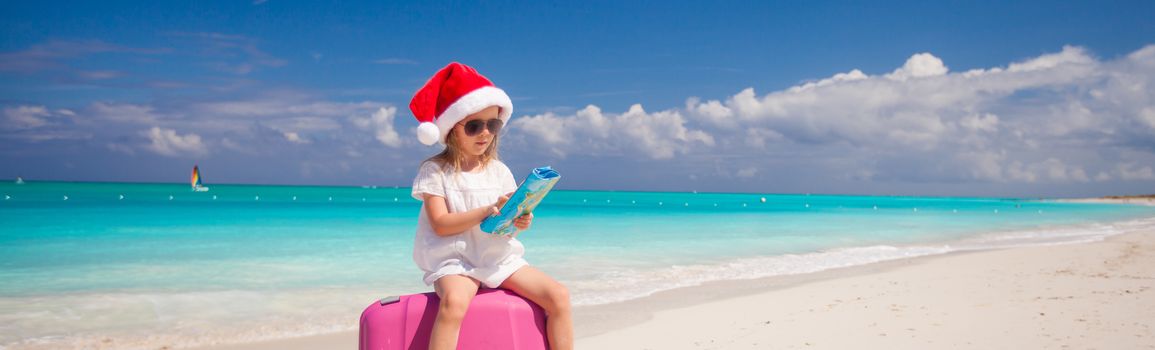 Little girl in Santa hat sitting on large suitcase at tropical beach