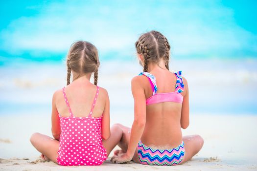 Adorable little girls playing with sand on the beach. Back view of kids sitting in shallow water and making a sandcastle