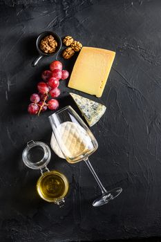 Cheese appetizer selection or whine snack set. Variety of mold french cheese, grapes, pecan nuts, white sauvignon wine and honey over black backdrop, top view, copy space vertical