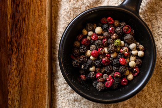 Mix of peppercorns in black bowl on linen cloth closeup near wooden board top view layflat