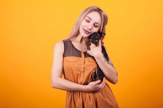 Beautiful young woman smiling and showing affection for her little puppy over yellow background