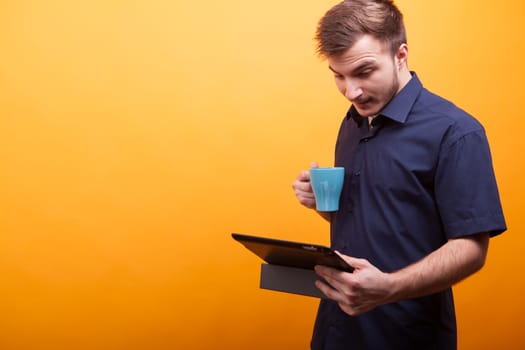 Young man in blue shirt drinking coffee and keeping tablet in hand over yellow background