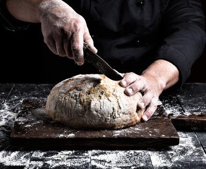 man cuts with a knife a round whole loaf of white wheat flour 