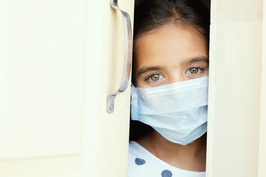 Young sad girl with medical mask wearing sneaking out through the home door - concept of home quarantine due to covid-19 or coronavirus outbreak.