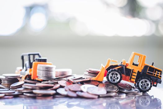 The yellow toy heavy machinery includes bulldozer and forklift with pile of coins against blurred background for saving money, investment, business and finance concept