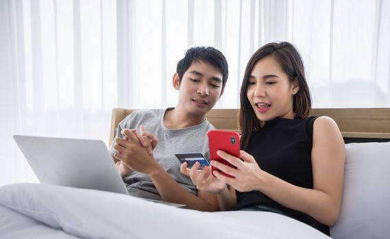 Asian Couple shopping online and paying with credit card on laptop and mobile phone at home,Happy couple at home surfing the net in bed