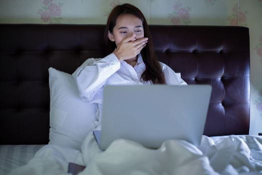 Asian woman using a notebook to work at night in bed and she yawns and sleepy.