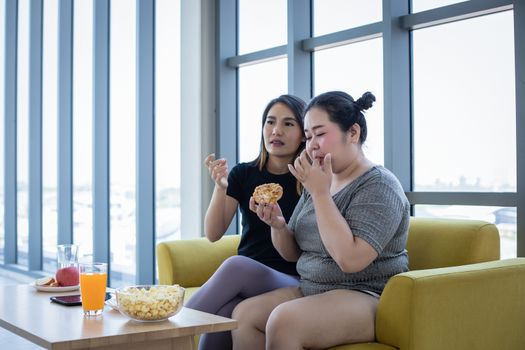 Overweight woman and asian girl enjoy eating food on sofa at hom