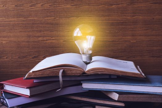 Open book with light bulb and hardback books on wood wall background. Selective focus