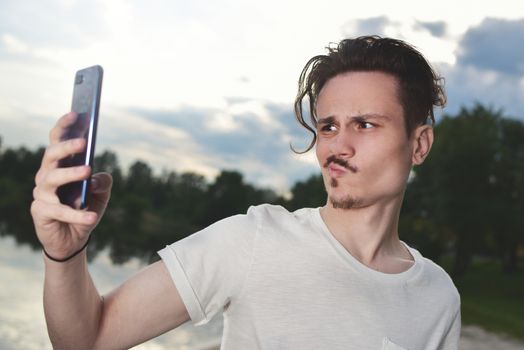 frustrated young handsome guy makes unsuccessful selfie on his smartphone in nature, the guy grimaces into the camera.the man read the bad news
