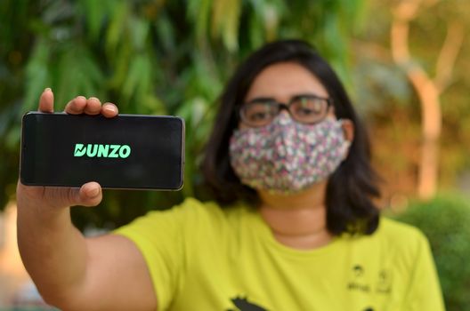 New Delhi, India, 2020. Girl wearing mask with Dunzo Delivery app logo glowing on the mobile phone screen in her hands. Selective focus and shallow depth of field. The main focus is on the logo.