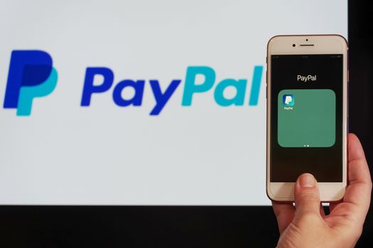 Hand holds before a screen mobile phone with PayPal application icon.